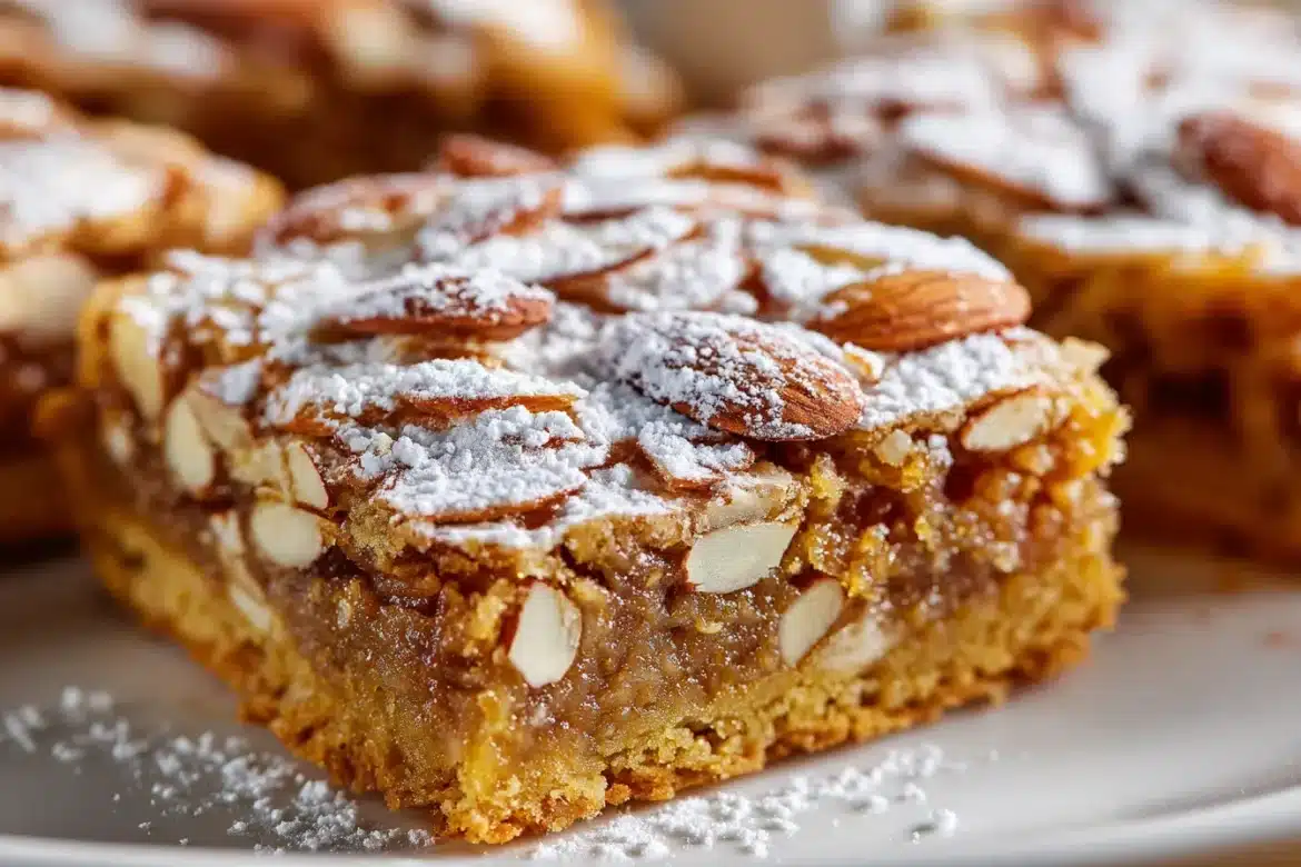Golden almond croissant cookie bars stacked on a white plate, with sliced almonds and powdered sugar on top, showing chewy interior.