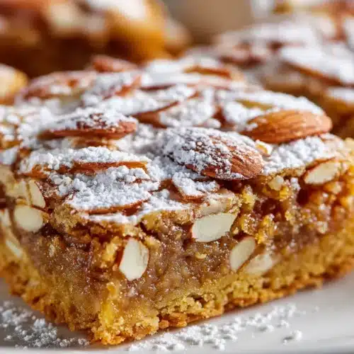 Golden almond croissant cookie bars stacked on a white plate, with sliced almonds and powdered sugar on top, showing chewy interior.